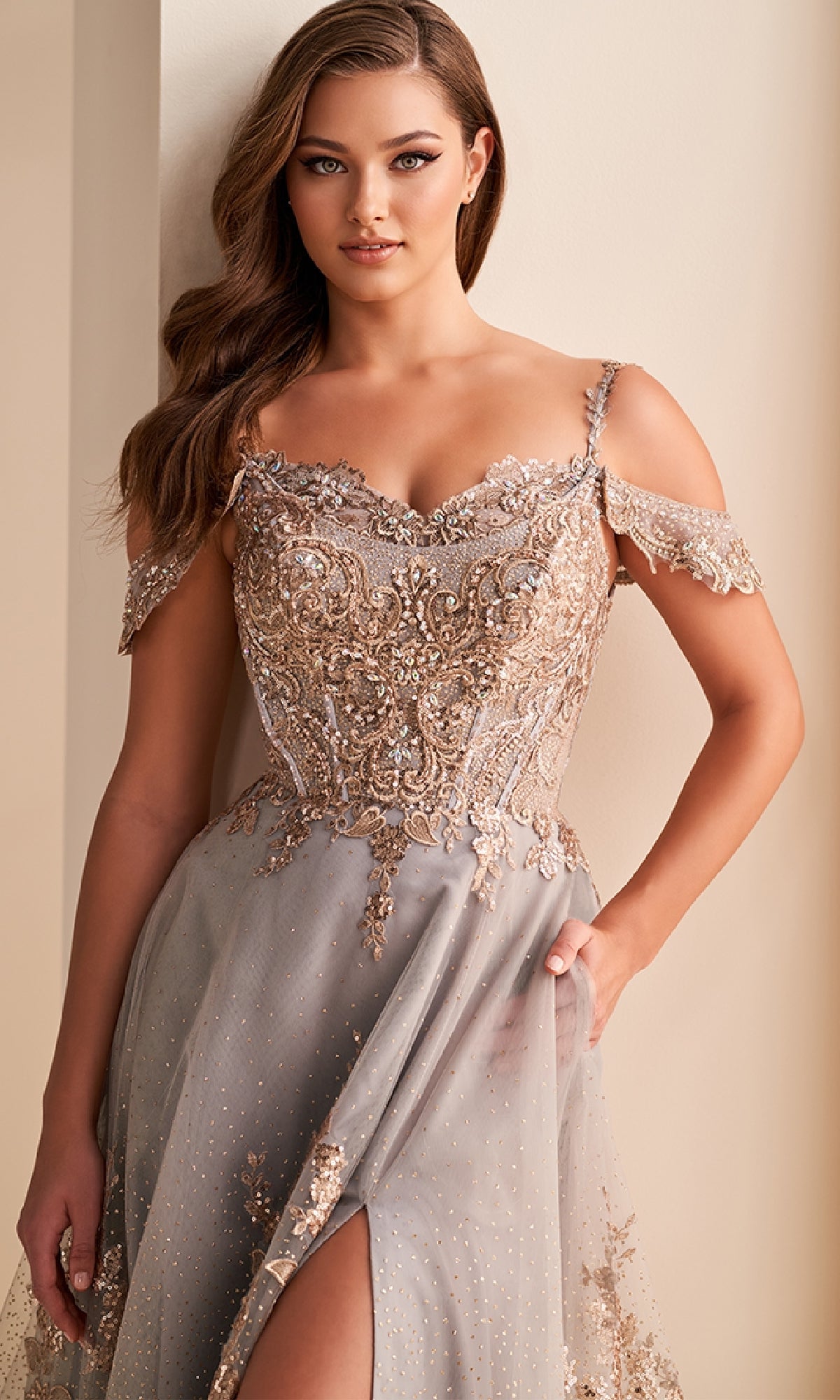 A woman stands against a wall wearing the Ellie Wilde EW36220 long prom dress, featuring light gray fabric, gold embroidery, off-the-shoulder straps, and a thigh-high slit.